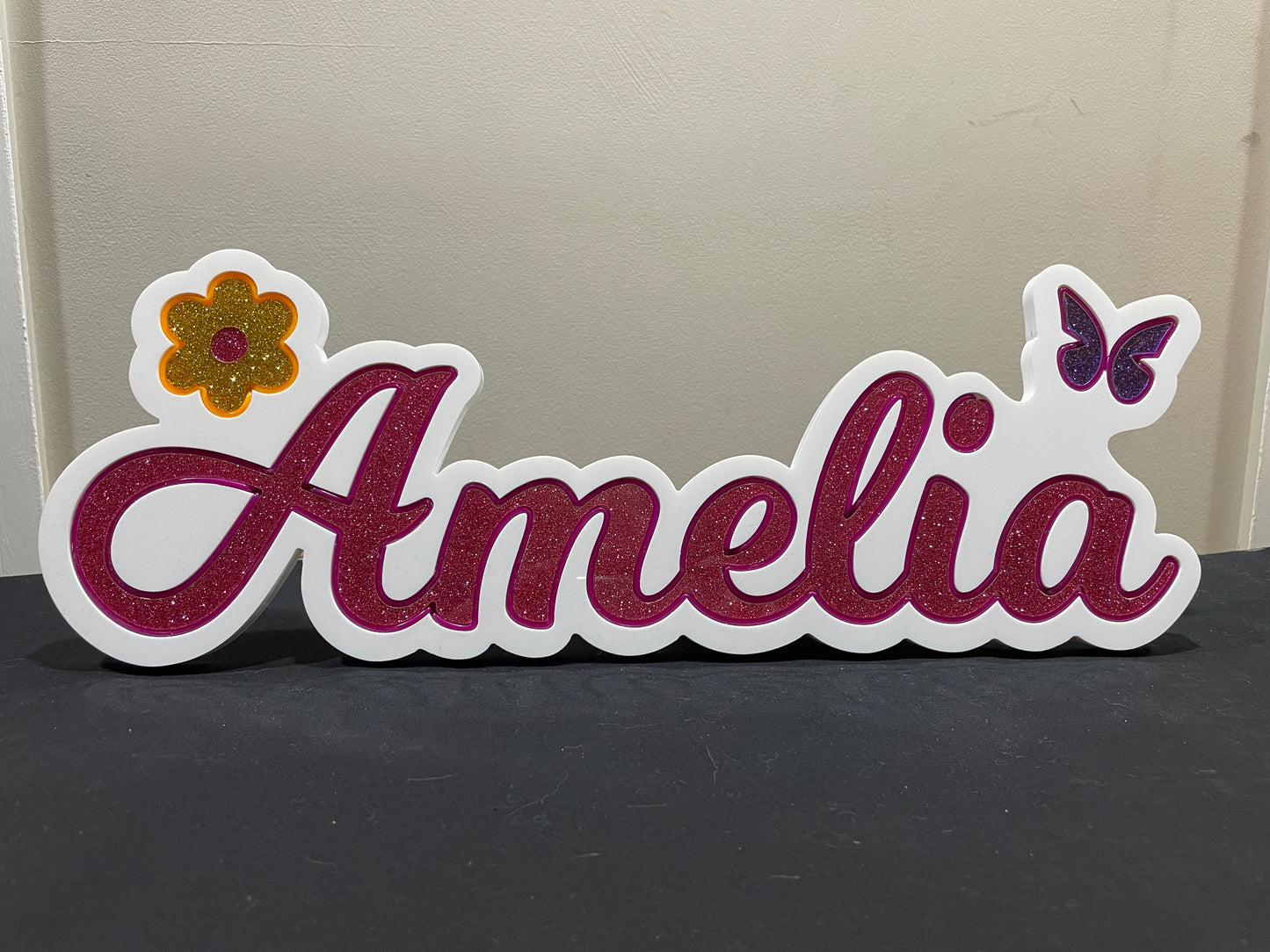 Amelia neon name light in pink with a gold flower at the beginning and a purple butterfly at the end displayed during the day