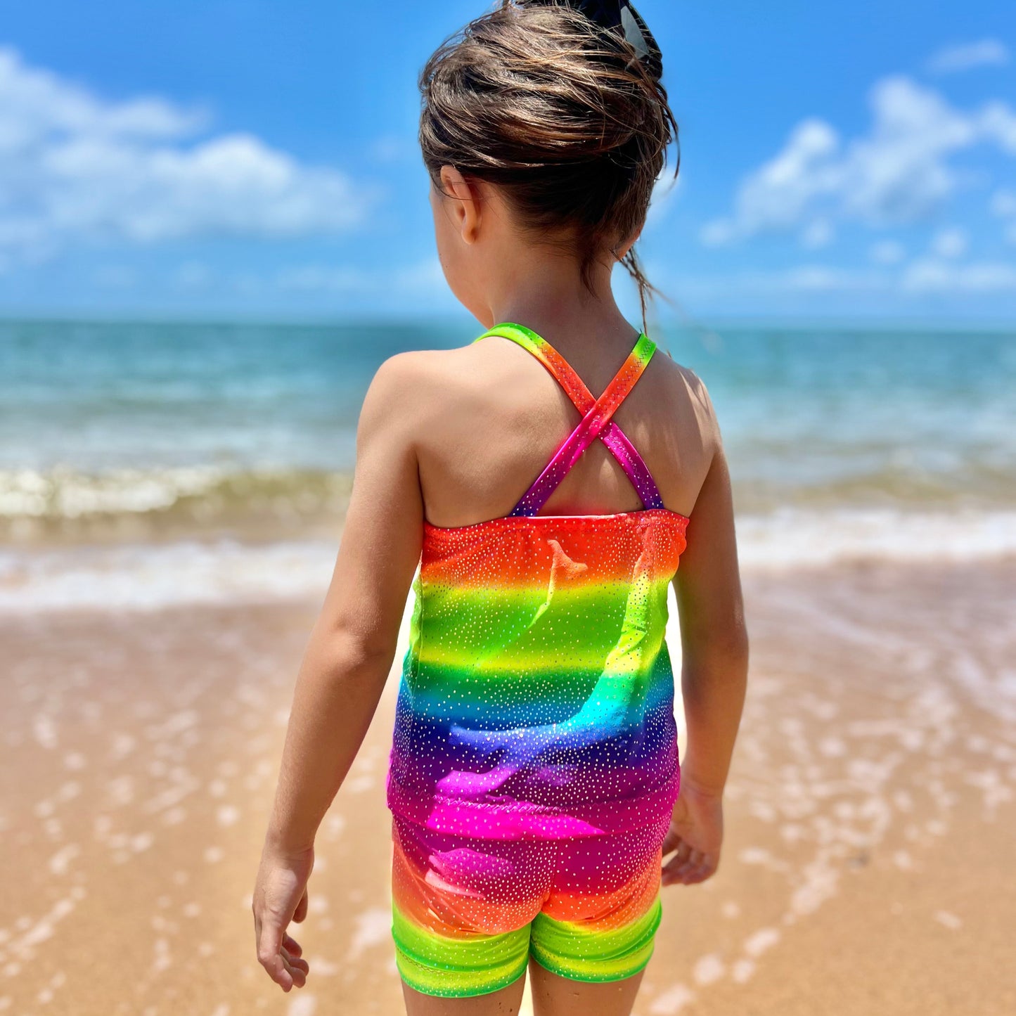 Back view of Girl wearing a bright rainbow crop top and shorts gymnastic outfit standing on a beach