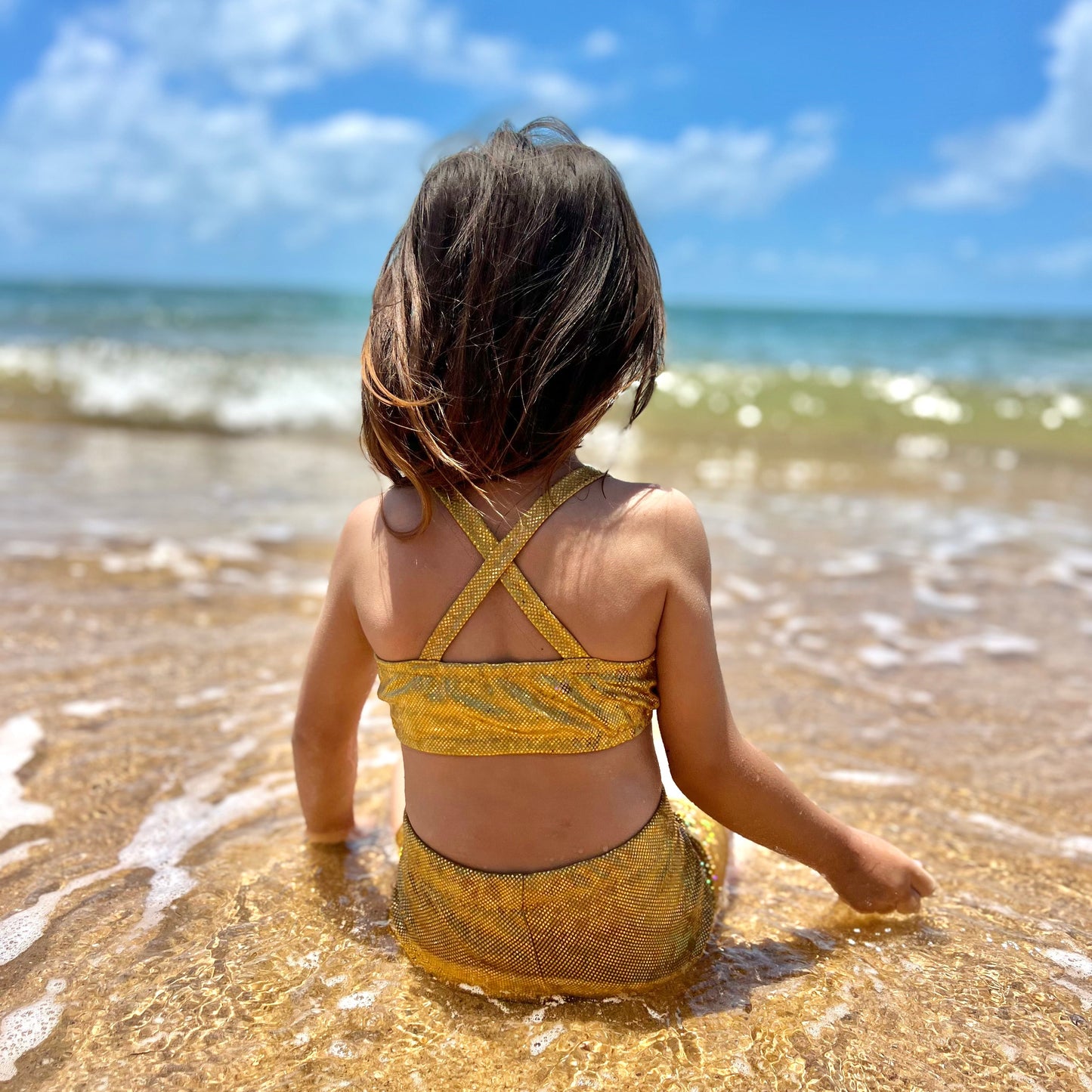 Back view of a Girl wearing a gold crop top and gold shorts gymnastic outfit siting on a beach