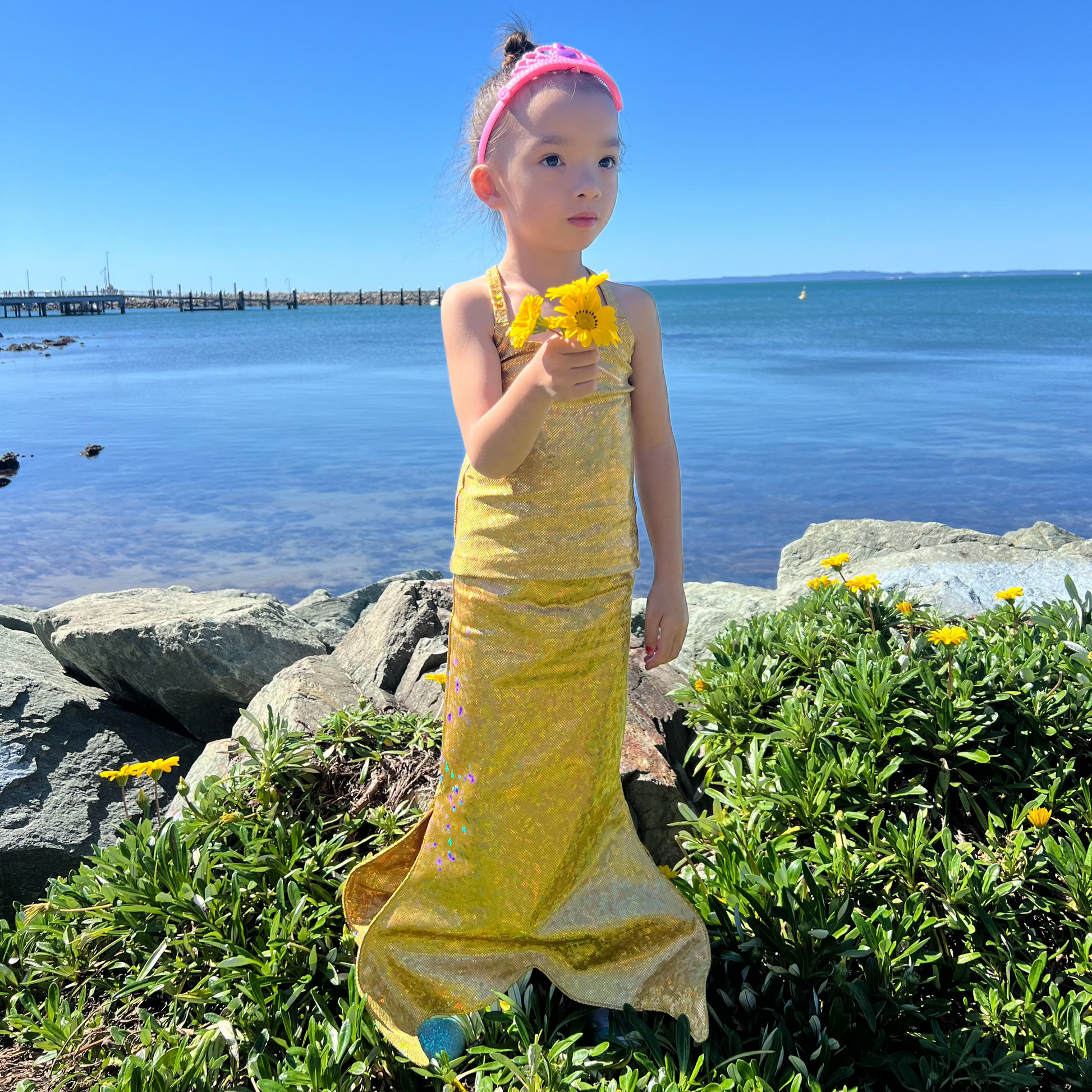 Girl standing near the ocean holding flowers wearing sparkly gold walkable dress-up mermaid tail costume and matching singlet top Brisbane Australia