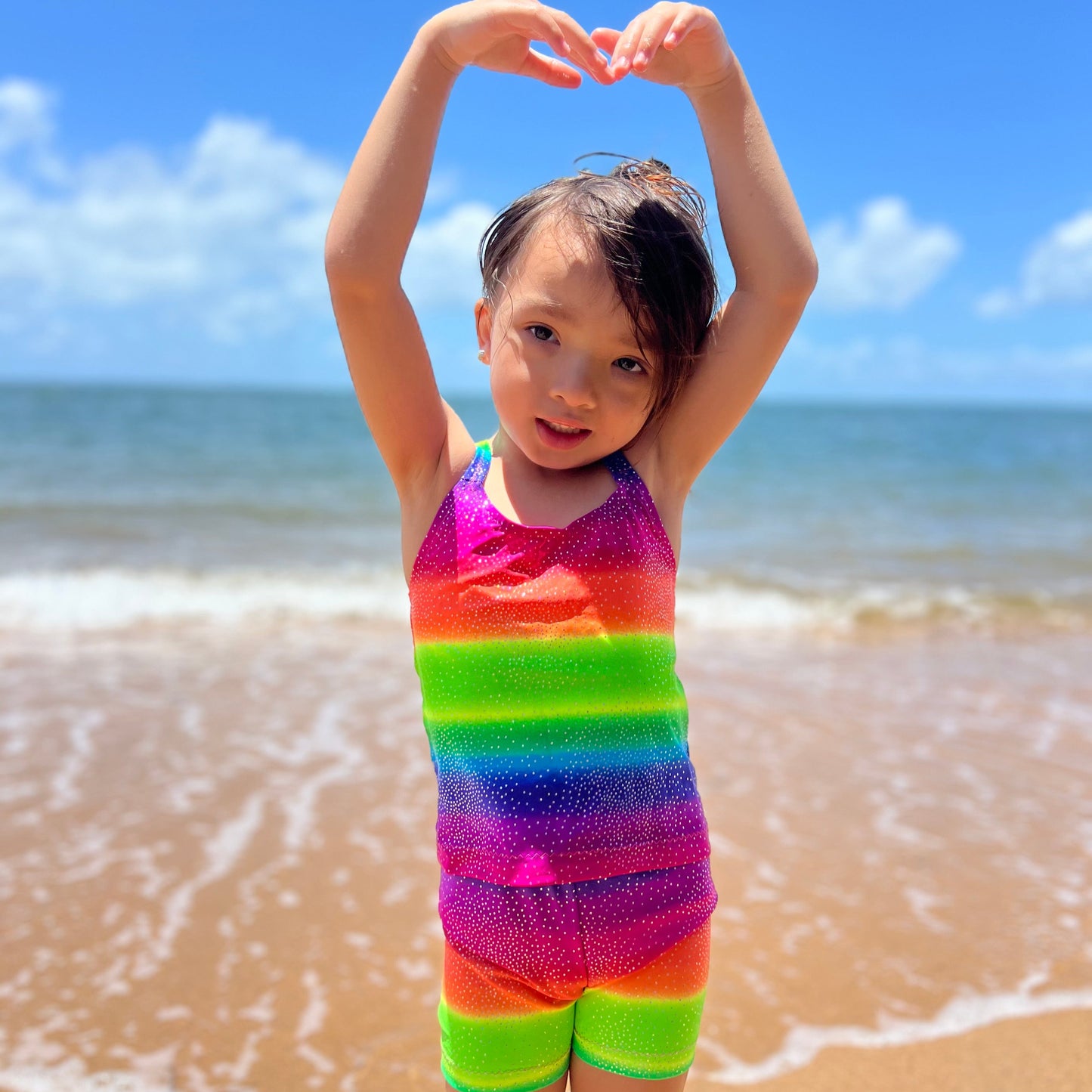 Girl wearing a bright rainbow singlet top and shorts gymnastic outfit on a beach with her hands above her head
