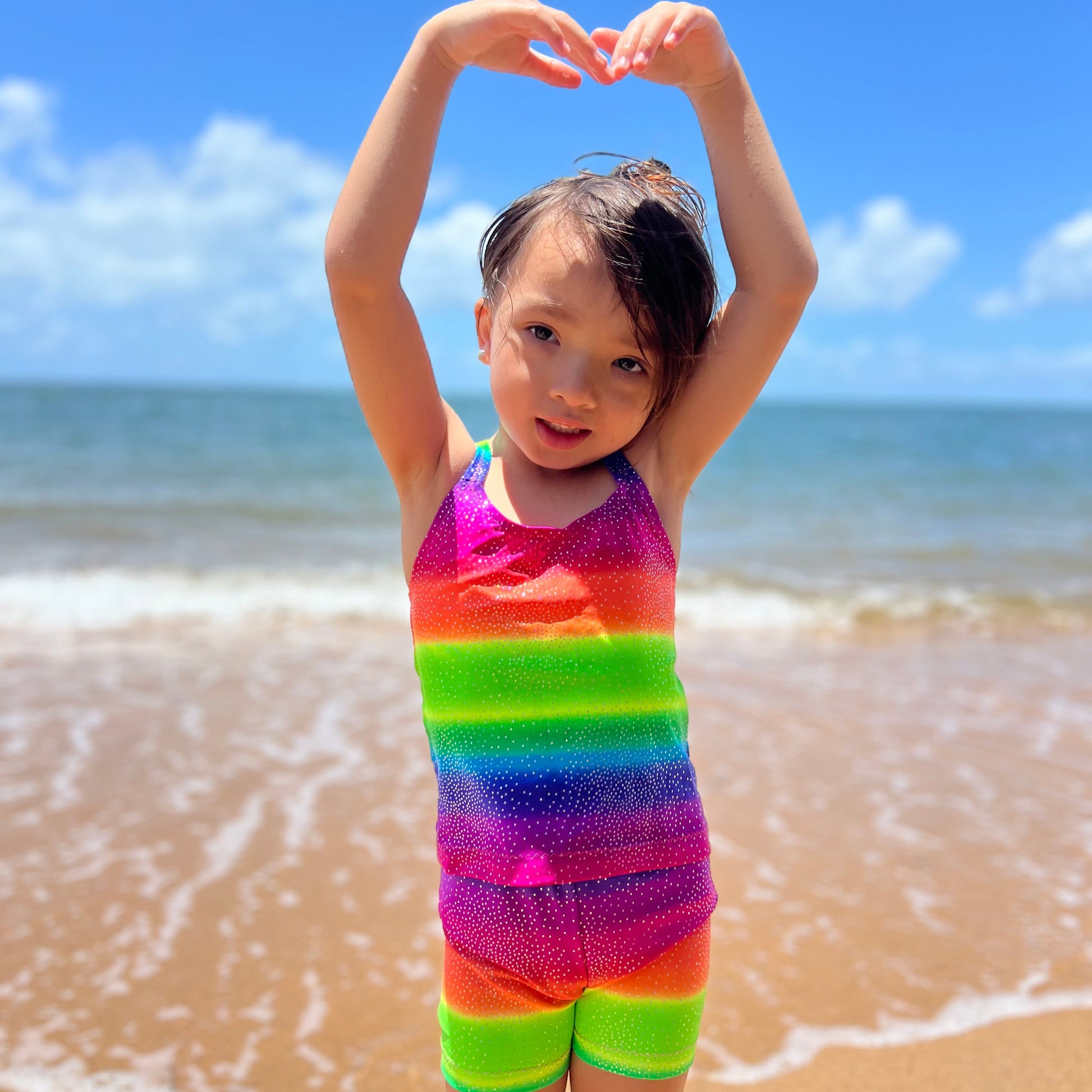 Girl wearing a bright rainbow singlet top and shorts gymnastic outfit on a beach with her hands above her head