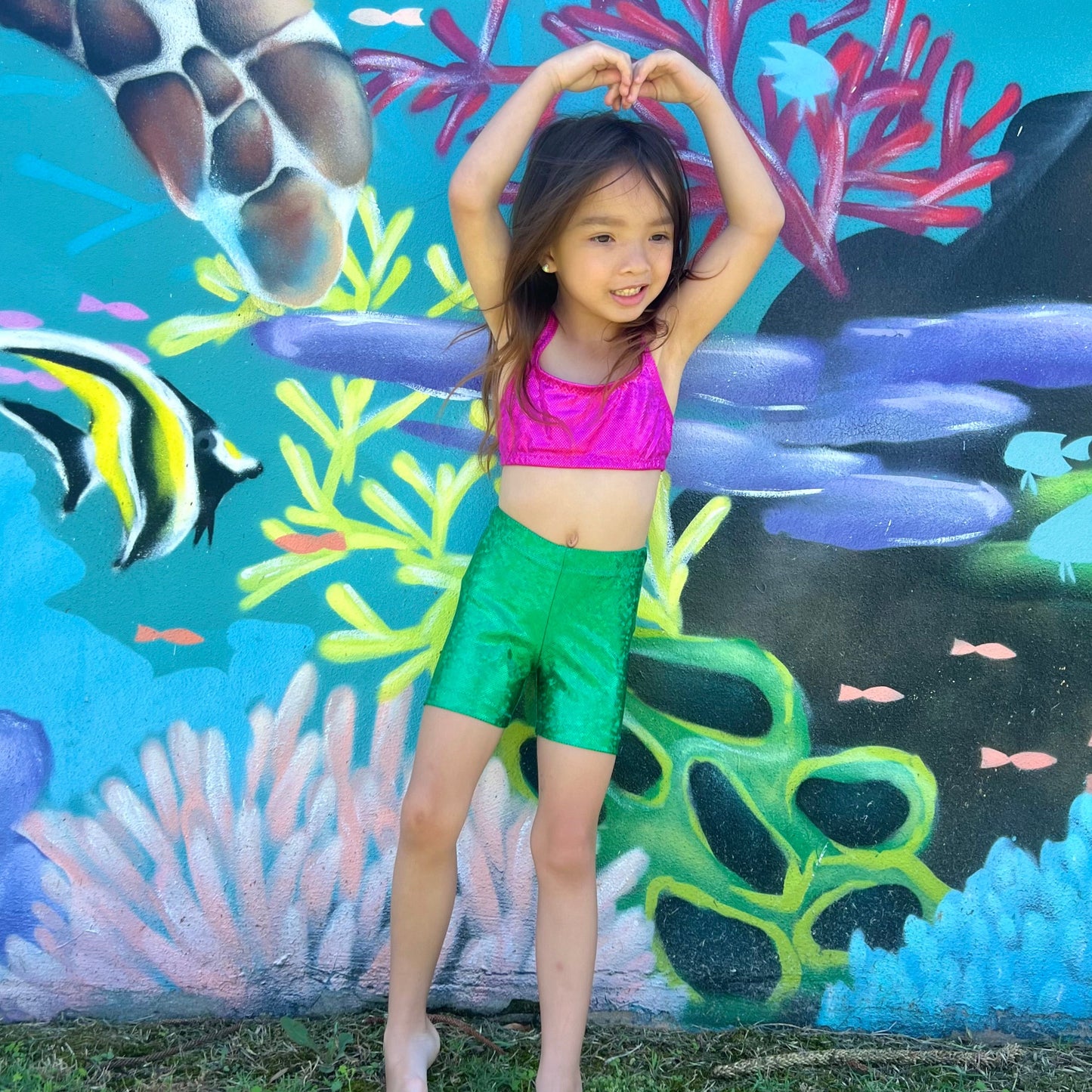 Girl wearing a hot pink crop top and green shorts gymnastic outfit with her hands above her head