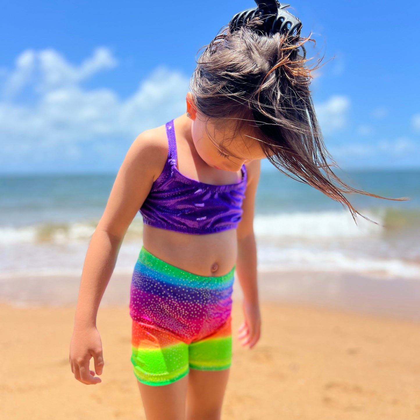 Girl wearing a purple crop top and bright rainbow shorts gymnastic outfit on a beach looking at the sand