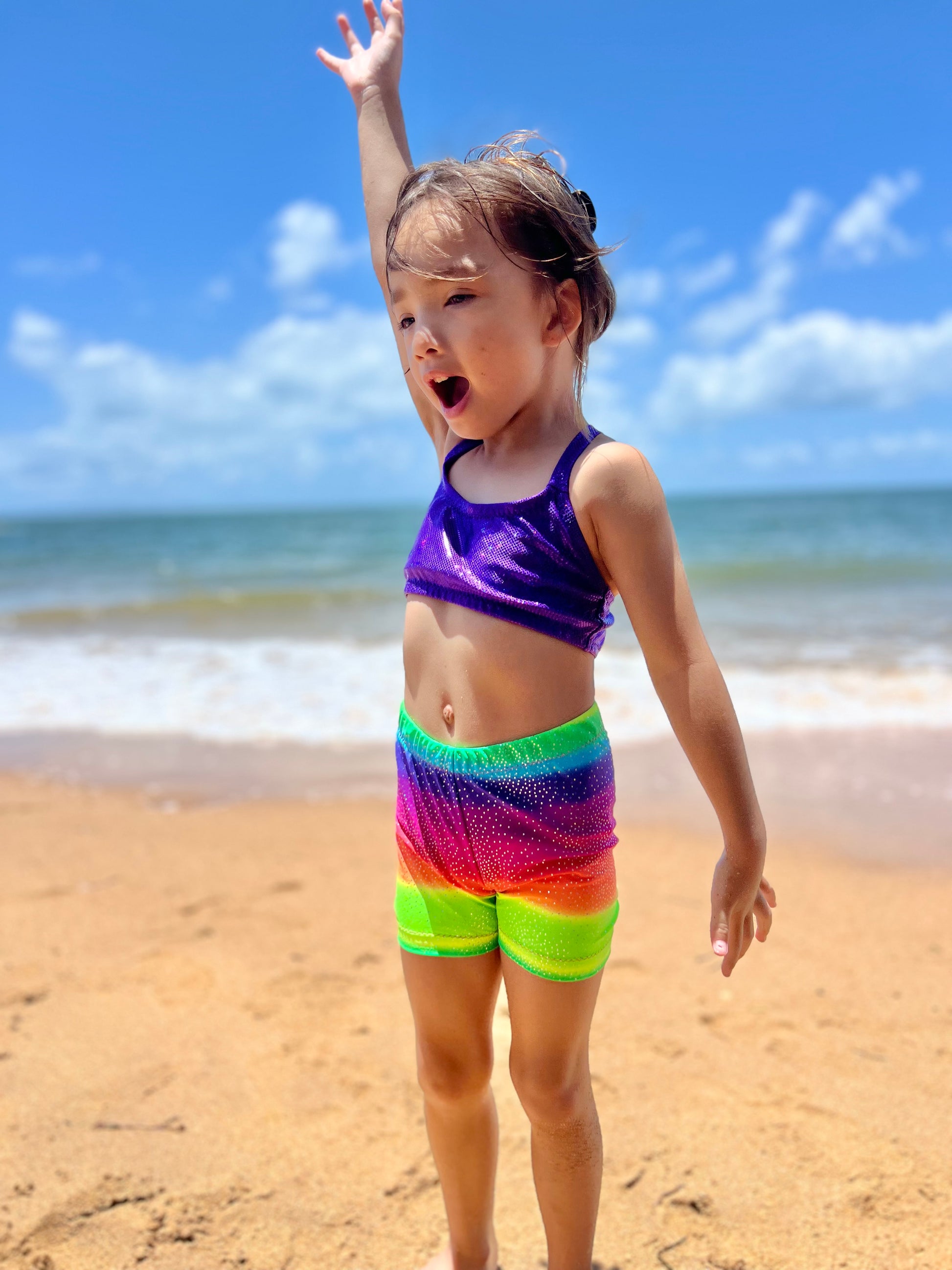 Girl wearing purple crop top and bright rainbow shorts on a beach with her hand in the air