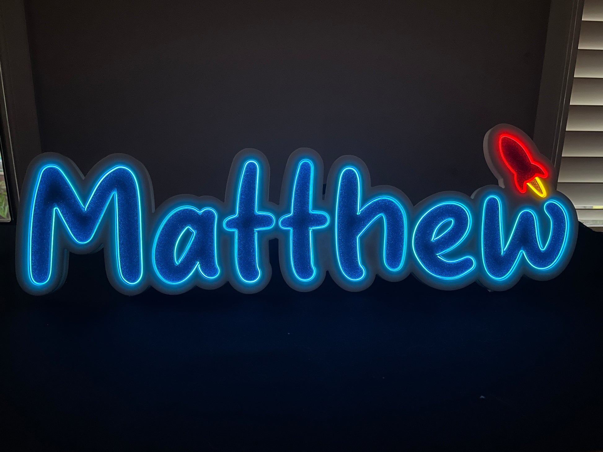 Matthew neon name light in blue and with a red rocket at the end
